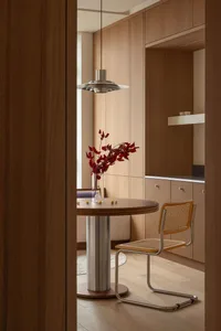 Modern minimalist breakfast nook featuring light walnut cabinetry, a round pedestal dining table, a cane and chrome chair, and a Louis Poulsen PH 5 pendant lamp with red floral arrangement.