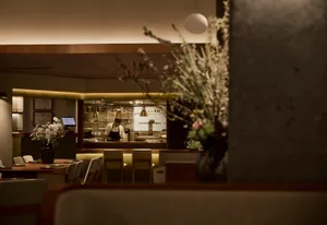 View from a dining table looking past a foreground vase of dried flowers toward an open kitchen where a chef is working under warm pendant lights, with wooden tables, upholstered chairs, and concrete walls.