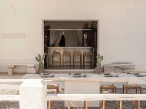 Outdoor dining area with a long travertine table, woven stools, potted cacti, and a view through a large opening to an indoor bar with wooden stools.