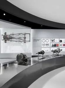 Modern exhibition hall with curved stainless steel counters displaying car engines, transmissions, and chassis components, with technical diagrams and informational panels on the white back wall.
