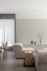Modern minimalist living room featuring a curved bouclé lounge chair, a travertine block side table, fluted wall paneling, and neutral beige tones.