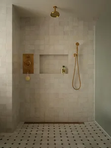 Minimalist shower space with off-white zellige tiled walls, a recessed niche holding a bath product, unlacquered brass shower head and mixer, and geometric black-and-white mosaic floor tiles.