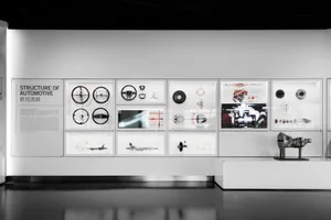 Museum exhibition wall displaying various automotive parts including steering wheels, driveshafts, brakes, and engine components in illuminated acrylic cases with technical diagrams and explanatory text.