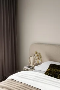 Modern minimalist bedroom corner featuring a textured beige fabric headboard, white bedding, an olive green velvet quilted pillow, and a marble nightstand with a sculptural white vase holding white orchids.