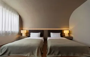 Modern minimalist bedroom with two single beds, dark fabric headboards, taupe bedding, and a distinctive curved plaster ceiling with warm ambient lighting.