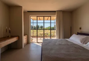 Modern minimalist bedroom featuring a large bed with grey bedding, wooden console desk, and floor-to-ceiling glass doors opening to a vineyard terrace.