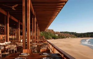 Open-air teak wood dining pavilion with wooden tables, woven chairs and ceiling fans, overlooking a tropical beach and ocean under clear blue sky.