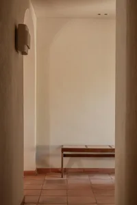 Minimalist entryway featuring terracotta floor tiles, a wooden bench with woven seat, and a sculptural wall sconce under an arched alcove.