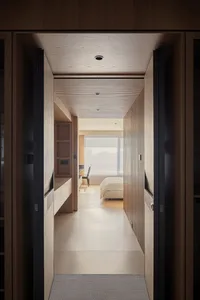 View from a doorway looking down a minimalist hallway with oak wood walls, built-in storage, and a wood slat ceiling leading to a bright bedroom with a large window.