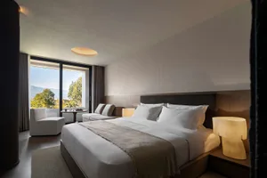 Modern hotel bedroom with large bed dressed in white linens, dark wood headboard, upholstered sofa, and large windows overlooking green mountains and trees.