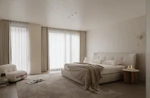 Minimalist bedroom with a large upholstered bed dressed in beige linen, a curved white bouclé sofa, sheer curtains allowing soft daylight, and a wooden side table with candles.