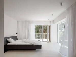 Modern minimalist bedroom with a dark wood floating platform bed, white bedding, sheer white curtains, and large glass doors opening to a balcony with greenery.