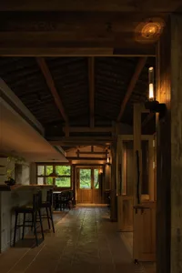 日式鄉村宅室內景觀，展現外露回收木天花板梁、大型石材地磚、混凝土廚房吧台搭配黑色木椅，以及在木牆上投射圓形光暈的溫暖壁燈。