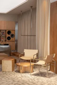 Modern living room featuring two cantilevered lounge chairs with chrome frames and cream upholstery, a round travertine coffee table, stone flooring, and a built-in wooden audio cabinet with speakers.