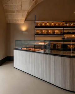 Modern bakery interior featuring a curved white fluted counter with glass display case, black marble top, and black metal shelving filled with packaged goods against a stone arched ceiling.