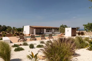 Modern white stucco villa with rectangular swimming pool, wooden deck, four lounge chairs, and xeriscape garden overlooking the sea under a clear blue sky.