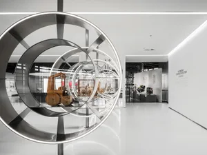Large circular stainless steel kinetic-style sculpture in foreground framing wooden vintage toy vehicles and modern car engines displayed in a white, brightly lit automotive technology exhibition space.