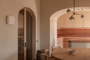 Warm minimalist kitchen and dining area featuring rounded wooden dining table, light oak cabinetry, terracotta marble backsplash, and dramatic arched doorways under a modern multi-arm pendant light.
