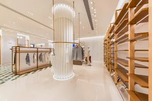 Modern minimalist clothing boutique interior with tall oak wood shelving units on the right, a large white pleated central column with integrated lighting, polished light-colored tile flooring, and clothing racks on the left.