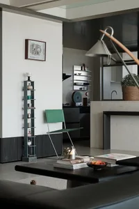 Modern minimalist living room featuring an emerald green chair, dark grey tall shelving unit, concrete kitchen island, and warm lighting from a sculptural floor lamp.