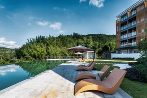 Modern woven rattan lounge chairs arranged on a stone terrace beside a reflecting pool, with a contemporary resort building and dense green bamboo forest in the background under a clear blue sky.