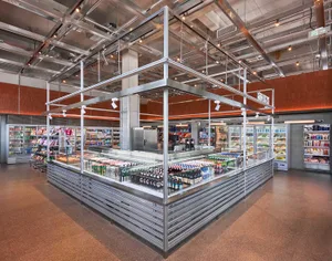Wide view of a modern supermarket featuring a large stainless steel central display island filled with beverages, surrounded by refrigerated cases and industrial metal ceiling structure.