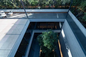 Bird's eye view of a modern concrete house featuring a central courtyard with a large tree, an elevated wooden deck with outdoor seating, and large glass windows.