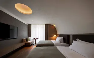 Modern minimalist twin hotel room with two beds, textured grey plaster walls, wooden flooring, built-in TV console, and warm pendant lighting.