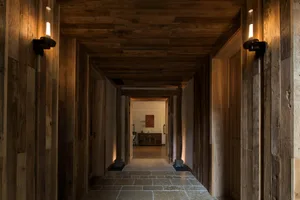 Long rustic hallway completely lined with reclaimed wood planks on walls, ceiling and doors, featuring black modern wall sconces casting warm light on the stone-tiled floor.