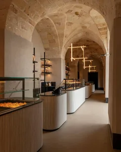 Interior view of a contemporary bakery housed within a historic stone-vaulted corridor, featuring curved fluted counters, glass display cases, and geometric pendant lights.