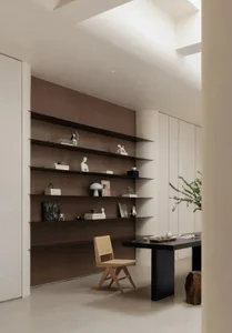 Modern minimalist home office featuring dark walnut built-in bookshelves, a wooden cane-back chair, black desk, and large skylight providing abundant natural light.