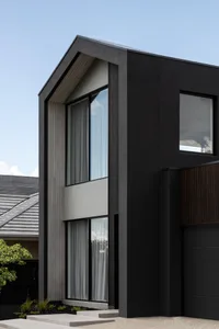 Modern two-story house exterior featuring charcoal black cladding, vertical light timber slats, large double-height windows with sheer curtains, and concrete steps under a clear blue sky.