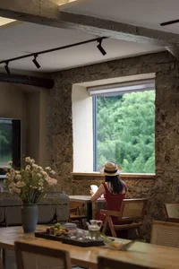 Woman sitting at a wooden dining table reading in front of a large window with a lush green forest view, surrounded by exposed stone walls and wooden furniture.