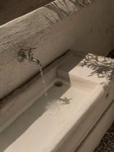 Close-up of a rustic outdoor utility sink made of travertine stone with a chrome faucet pouring water, casting strong leaf-shaped shadows on the surface.