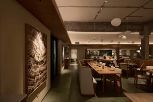 Modern Japanese restaurant interior with warm wood dining tables, upholstered banquettes, terrazzo flooring, exposed concrete beams, and large landscape artwork on the wall.