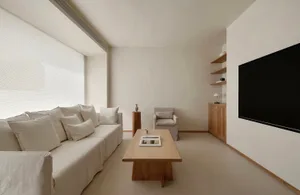 Minimalist living room with white L-shaped sofa, light oak coffee table, single armchair, and wall-mounted TV in a bright, neutral-toned space.