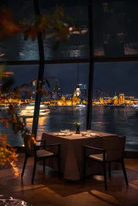 Elegant dining table set for two with dark wood chairs, positioned beside massive windows revealing the glittering nighttime skyline of Shanghai and the Huangpu River.