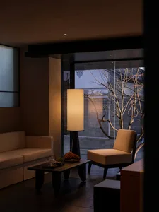 Modern minimalist living room featuring a beige sofa, single lounge chair, dark walnut coffee table, and a tall cylindrical floor lamp with warm glow, overlooking a courtyard with bare trees at dusk.