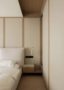 Modern minimalist bedroom corner showing a beige upholstered bed with white linens, a wall-mounted oak floating nightstand, and a slim white pendant light hanging from a wood-paneled ceiling.