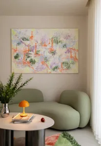 Sage green curved boucle sofa in a bright room corner, orange mushroom lamp on a round marble coffee table, large colorful abstract painting on the wall, and a silver vase with green foliage.
