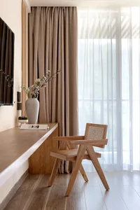 Minimalist wooden armchair with rattan seat and backrest placed next to a built-in oak desk and large window with sheer curtains
