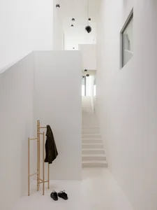 Minimalist white double-height entryway featuring a straight staircase, wooden coat rack with dark green jacket, black shoes on floor, and multiple black spherical pendant lights hanging from high ceiling.