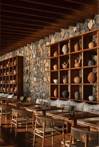 Rustic restaurant interior with dark wood ceiling beams, textured stone wall, and extensive wooden shelving displaying numerous terracotta and ceramic pottery pieces, paired with wooden dining tables and woven rattan chairs with light cushions.