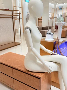 White articulated female mannequin seated on a burl wood chest of drawers in a bright, modern retail boutique with neutral tones and designer accessories.