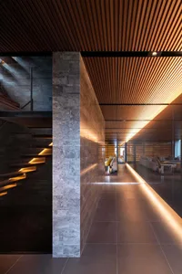 Modern luxury hallway featuring grey marble walls and floors, dark wood slat ceiling with integrated LED lighting, floating staircase with underlit steps, and a view into a living area with wine cellar in the background.