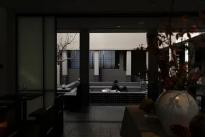 Man sitting in a modern sunken concrete courtyard lounge inside a minimalist Japanese-style house, viewed from a dark dining area with foreground vase.