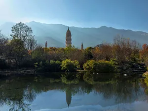 Three ancient pagodas reflected in the calm water of a lake surrounded by autumn trees and distant mountains under a clear blue sky.