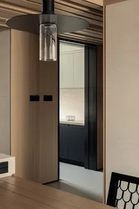 Modern minimalist dining area looking through a black-framed opening into a kitchen with warm oak cabinetry and matte black lower cabinets under soft lighting.