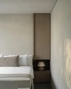 Modern minimalist bedroom featuring a bed with white and beige linens, built-in oak wardrobe, and a small ceramic table lamp on a wooden nightstand.