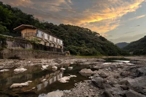 現代混凝土河畔建築搭配大面窗，俯瞰淺水多岩石的河流，背景為茂密綠意山區，拍攝於日落時分。